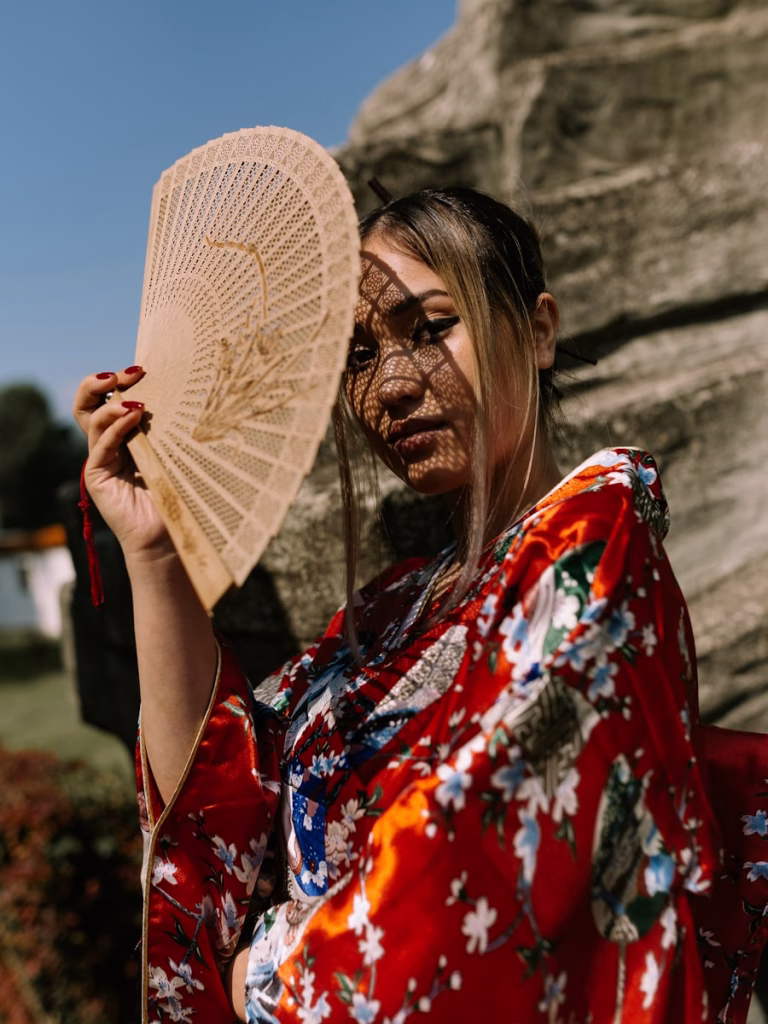 woman in red, white, and blue floral kimono standing and covering her face with wooden hand fan